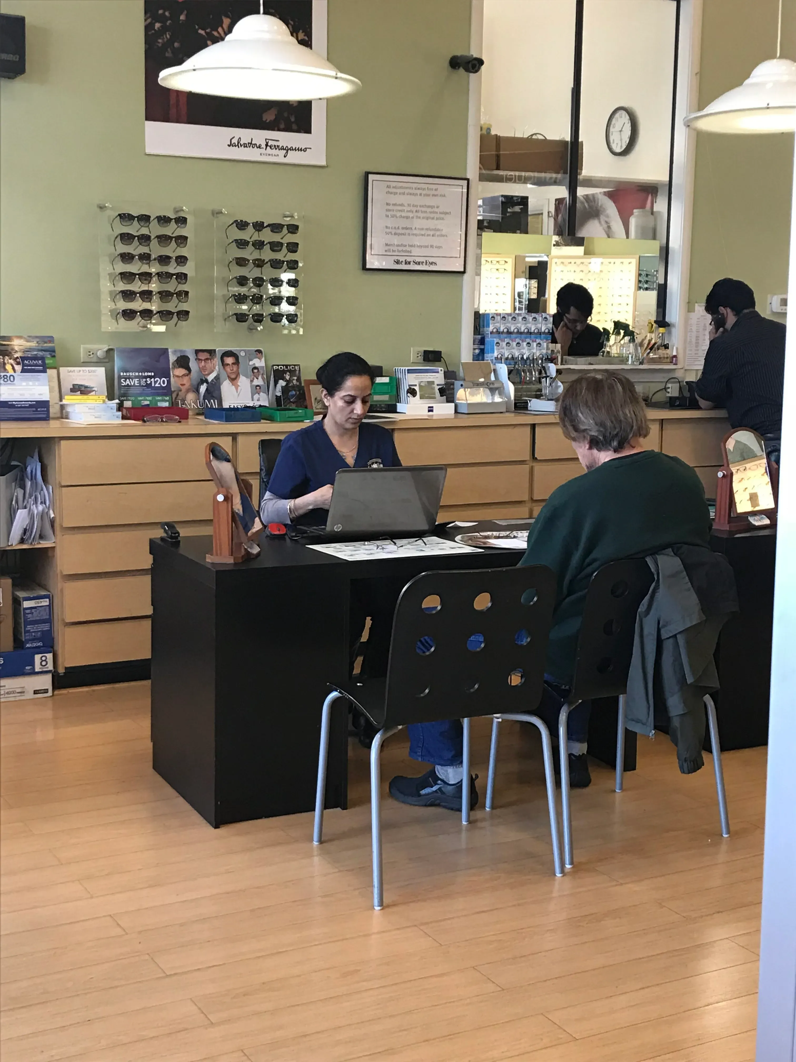 Inside Site for Sore Eyes - Fremont, a person consults with an employee at a desk, surrounded by shelves with eyewear and optical equipment.